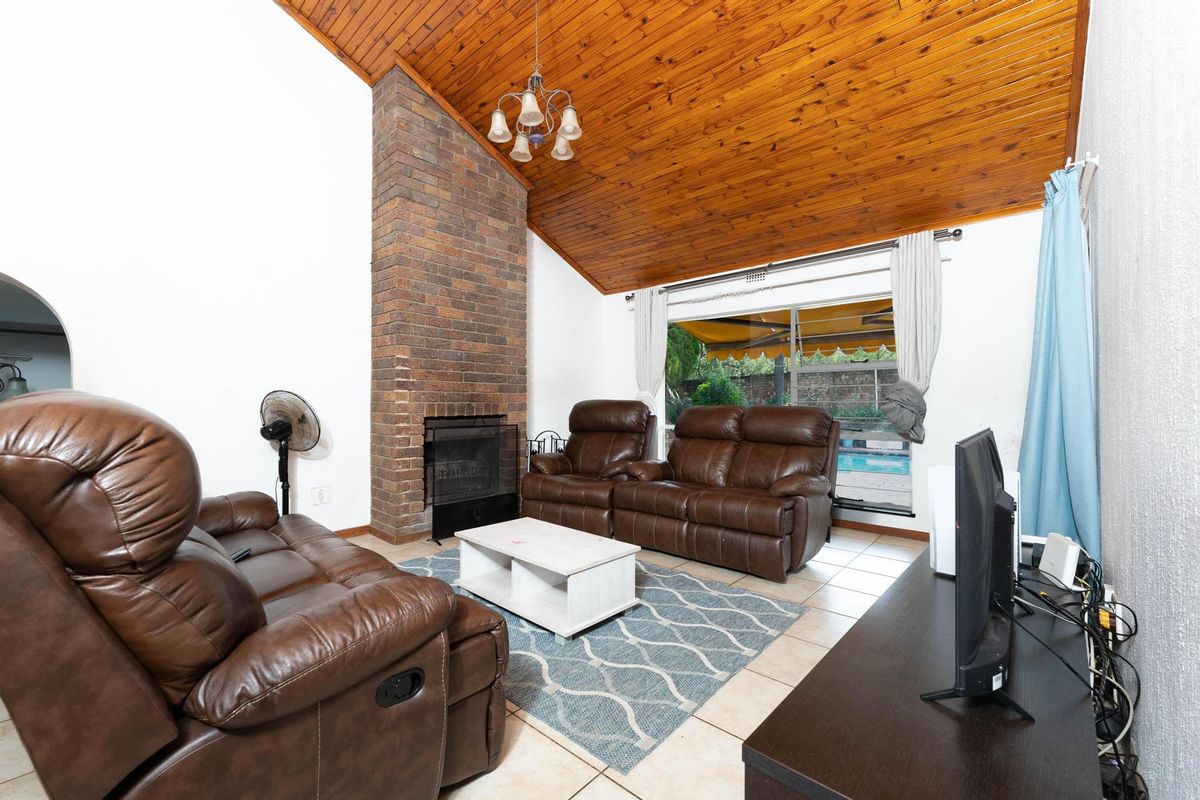 Spacious living room featuring a prominent brick fireplace, vaulted timber ceiling, and tiled flooring with direct access to the outdoor pool area.