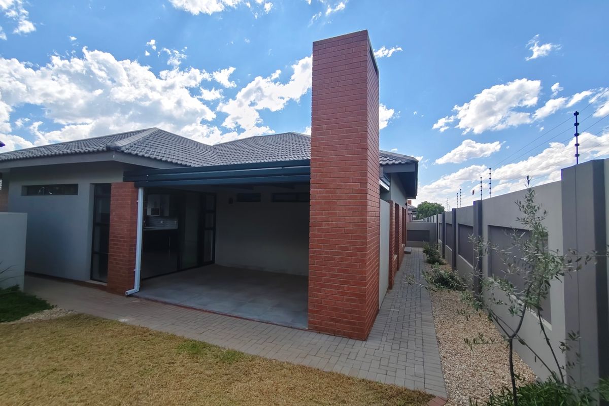 A well-maintained rear exterior view of the townhouse featuring a covered patio, brick chimney, paved pathways, and secure boundary wall with electric fencing.