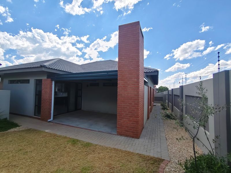 A well-maintained rear exterior view of the townhouse featuring a covered patio, brick chimney, paved pathways, and secure boundary wall with electric fencing.