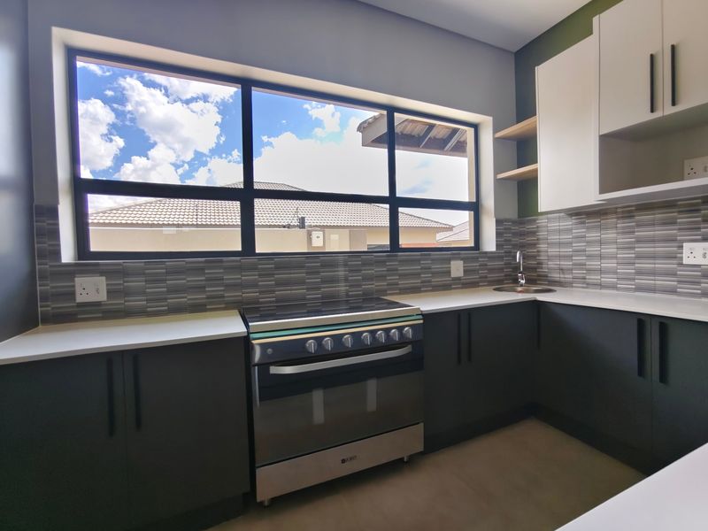 Modern kitchen featuring dark cabinetry, stone countertops, a stainless steel stove, and a tiled backsplash with a large window providing natural light.