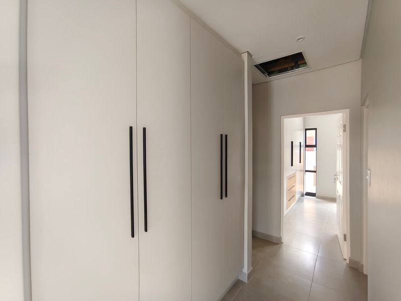 A bright, modern hallway featuring extensive built-in cabinetry, neutral tiled flooring, and clean finishes, leading towards a secondary passage area.