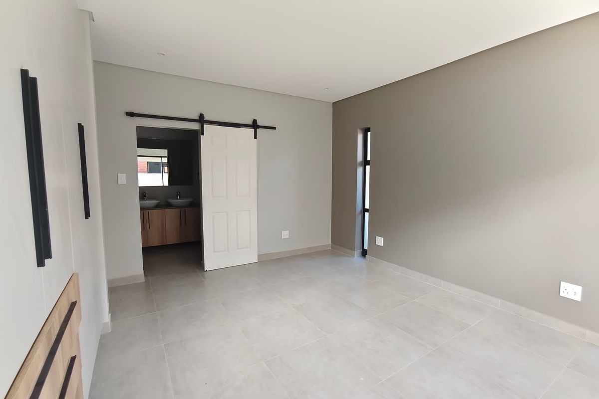 A modern, empty bedroom featuring neutral tones, tiled flooring, a sliding barn door, and an adjoining bathroom with dual vanity.