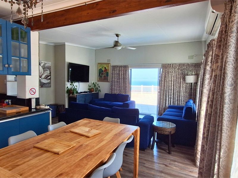 Spacious open-plan living, dining, and kitchen area featuring exposed timber beams, modern blue cabinetry, and direct access to an outdoor patio with ocean views.