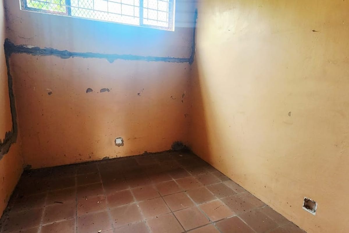 An empty bedroom featuring tiled flooring and security bars on the window, currently requiring wall repairs and electrical finishing.