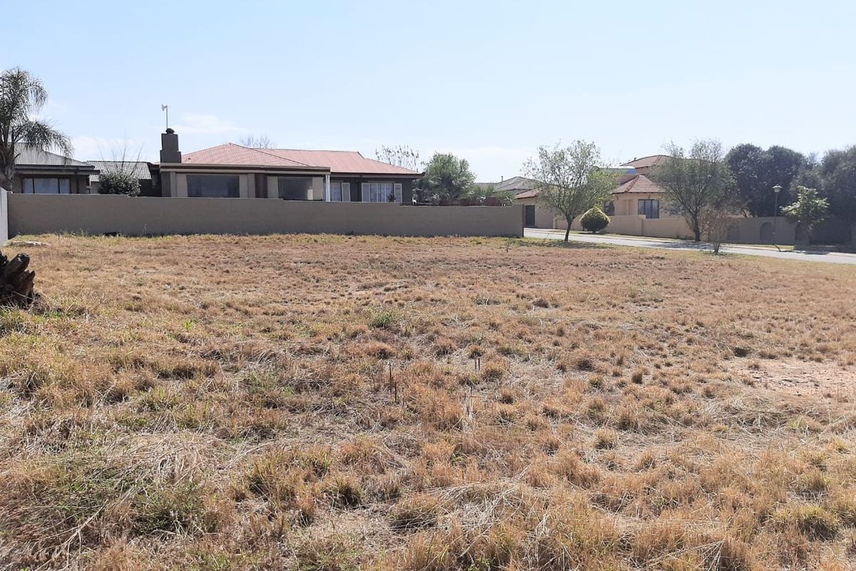A vacant residential plot featuring dry grass, enclosed by a boundary wall, situated in a developed suburban neighborhood.