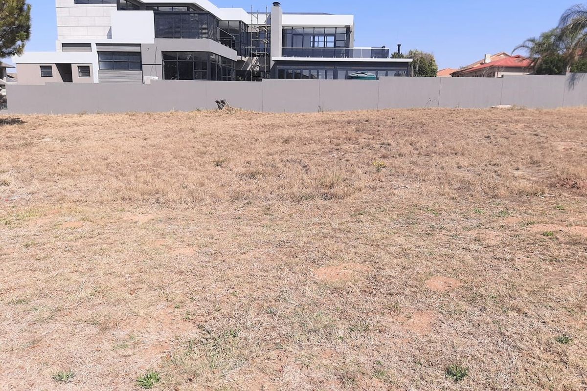 A view of a vacant residential plot of land with a boundary wall and neighboring modern property in the background.
