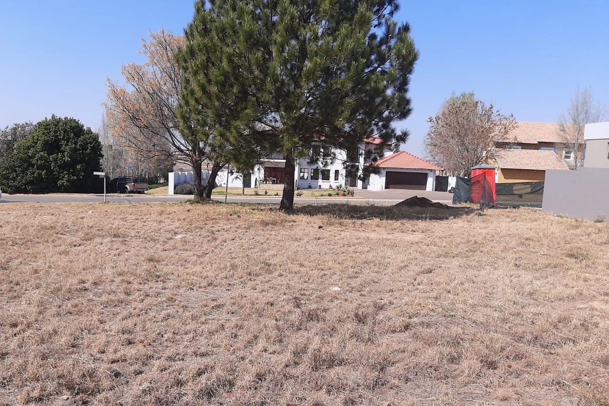 A vacant residential plot featuring natural grassland and mature trees, offering a clear view of the surrounding neighborhood and street access.