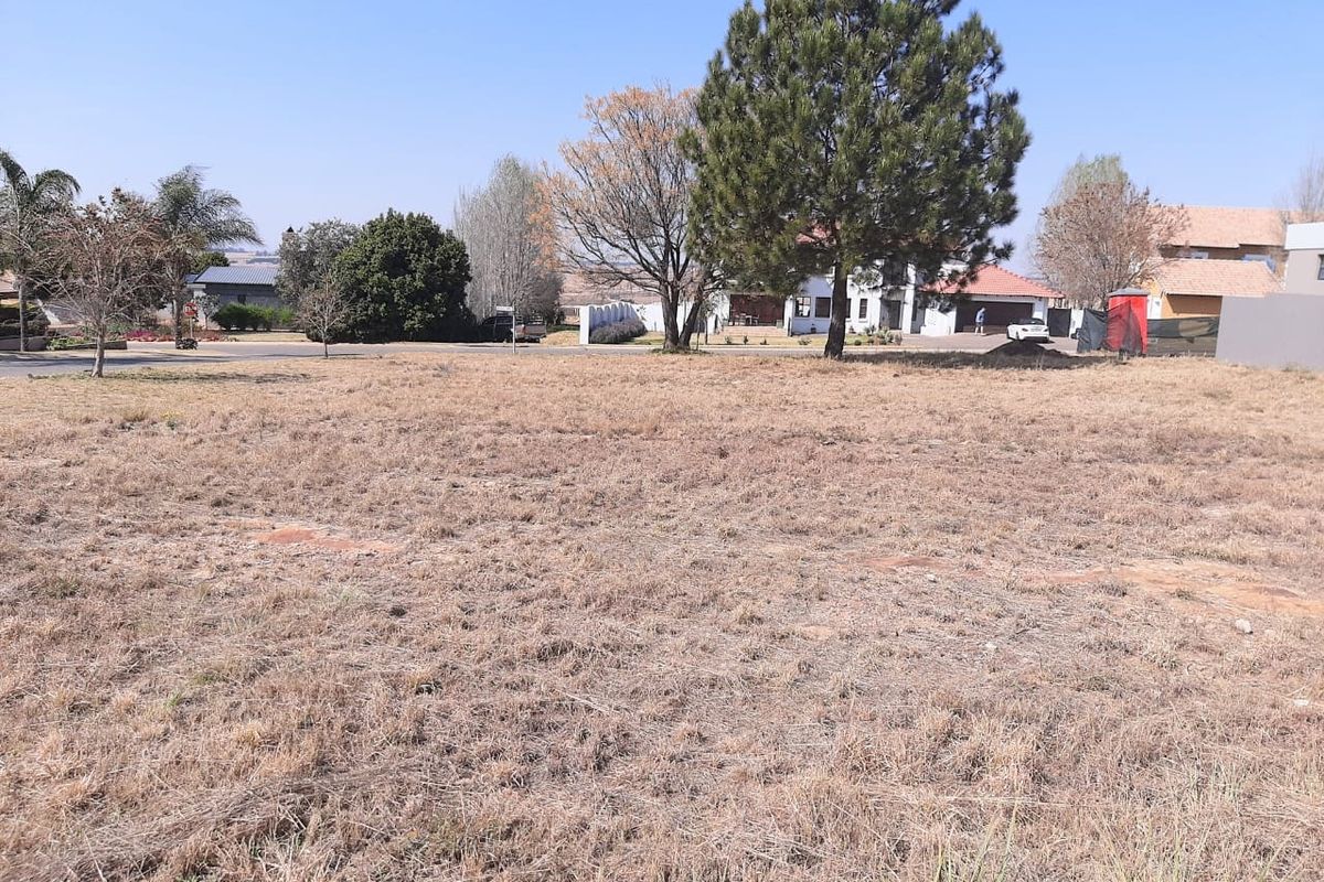 A view of a vacant residential plot featuring dry grass and mature trees, situated in a developed neighborhood.