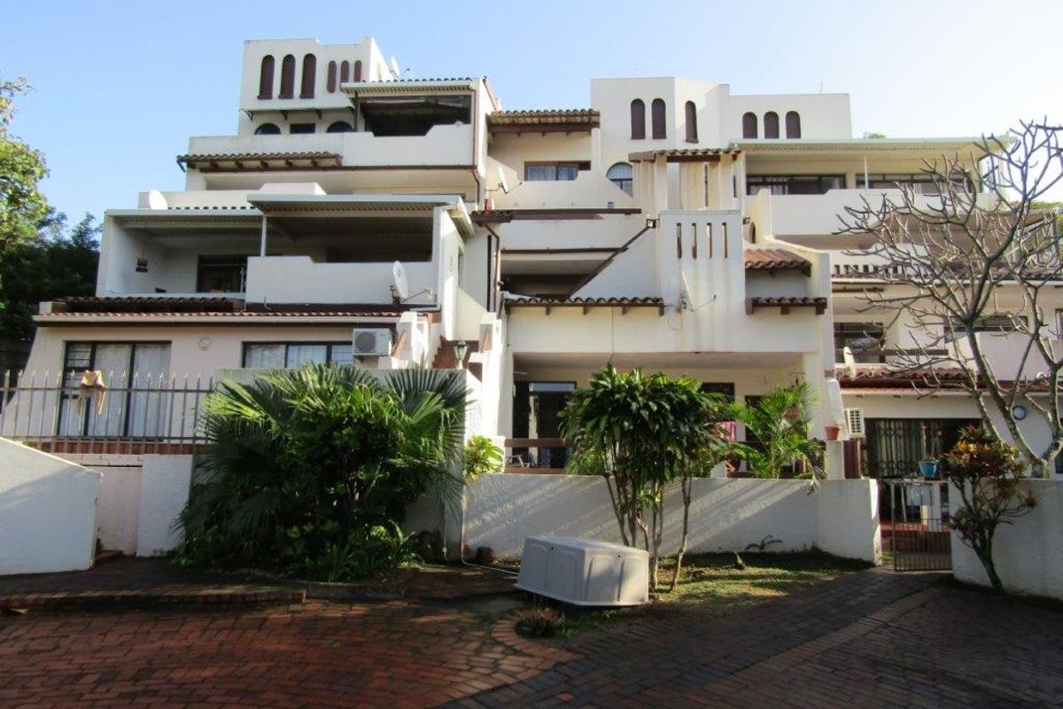 Rear elevation of a multi-story apartment complex featuring tiered balconies, paved driveway access, and established garden landscaping.