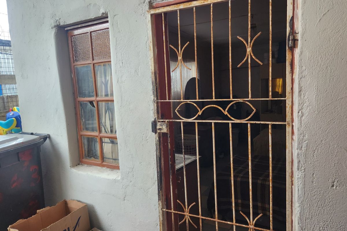 Exterior view of a rear entrance featuring a rusted security gate and a timber frame window set into a plastered masonry wall.