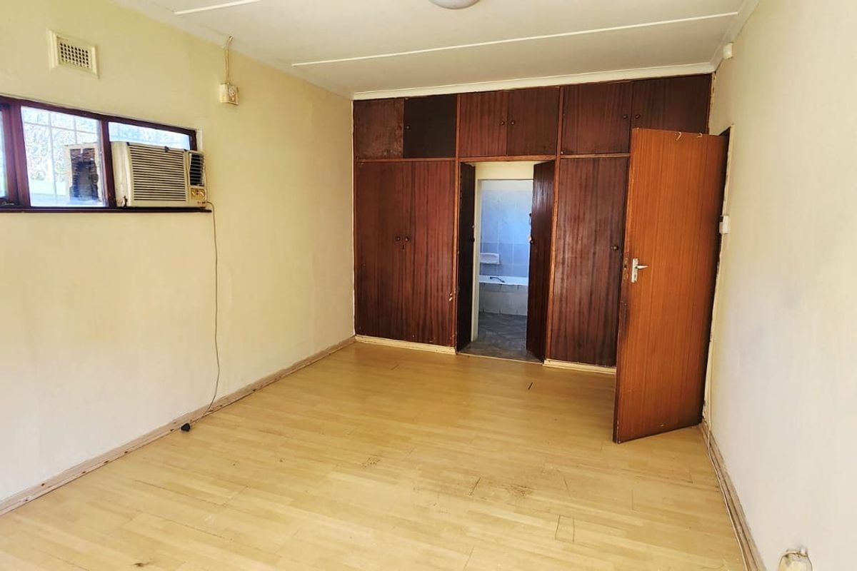 An empty bedroom featuring laminate flooring, built-in wooden cupboards, and a window-mounted air conditioning unit, requiring cosmetic updates and electrical maintenance.