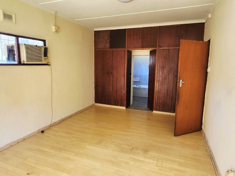An empty bedroom featuring laminate flooring, built-in wooden cupboards, and a window-mounted air conditioning unit, requiring cosmetic updates and electrical maintenance.