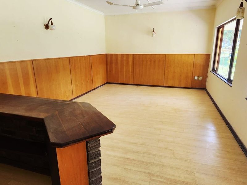 Spacious living area featuring a built-in bar, wood wall paneling, ceiling fan, and large windows with security bars, offering a blank canvas for renovation.