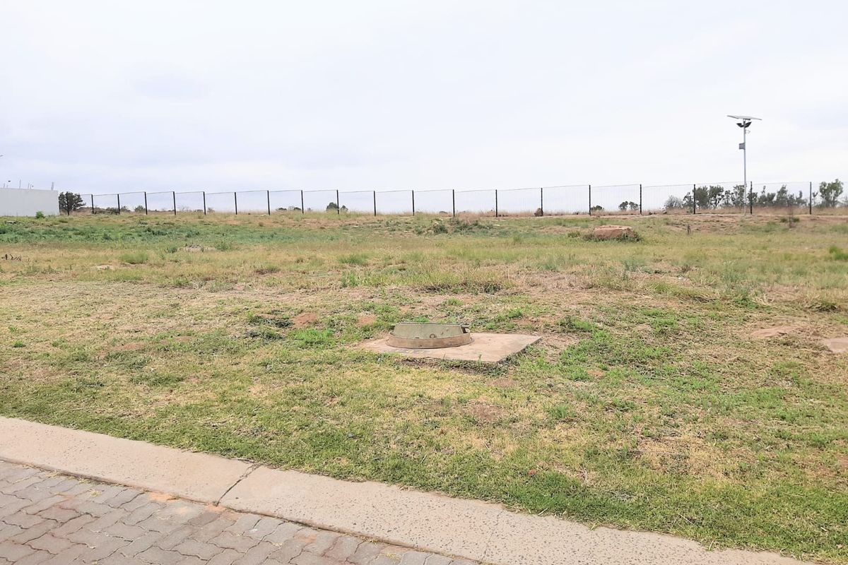 A vacant residential plot featuring a paved verge, perimeter fencing, and a central manhole cover, ready for development.