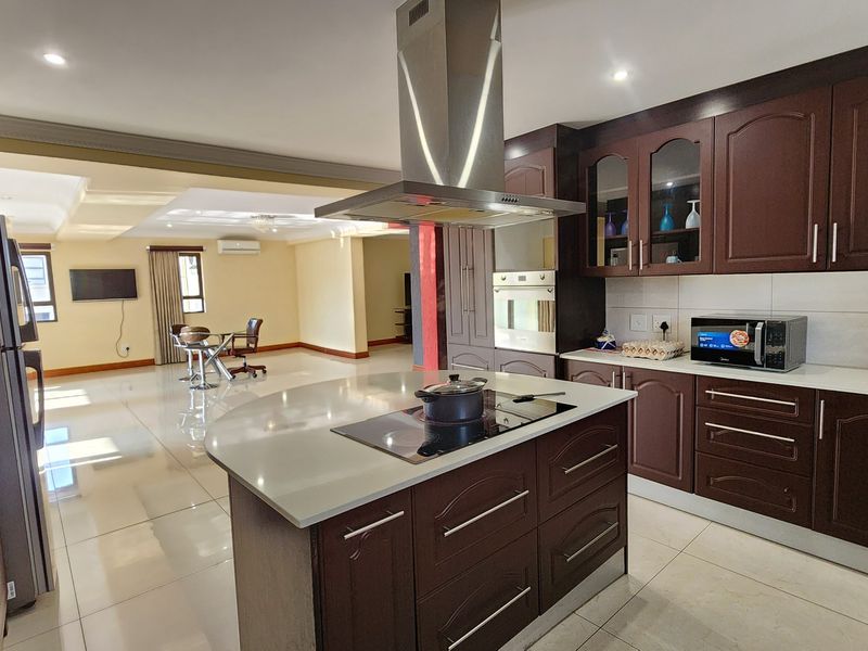 Spacious open-plan kitchen featuring dark wood cabinetry, a central island with cooktop, and integrated appliances, flowing into a bright living area.