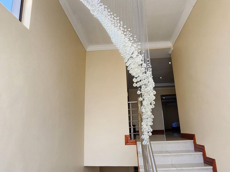 A bright, modern hallway featuring a grand spiral crystal chandelier, tiled staircase with stainless steel balustrade, and clean neutral wall finishes.