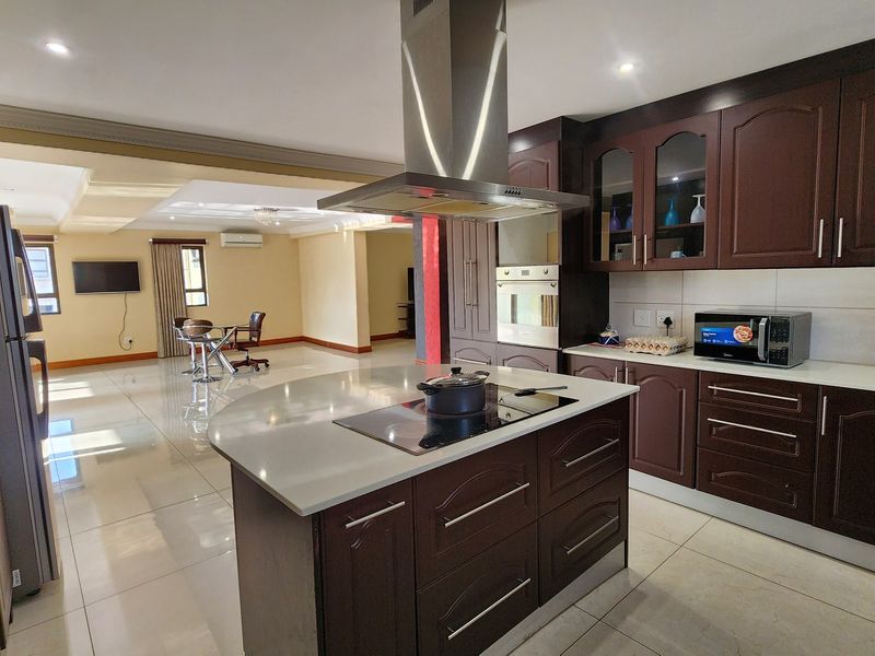 Spacious open-plan kitchen featuring a central island with cooktop, dark wood cabinetry, and integrated appliances, flowing into a bright living area with tiled floors.