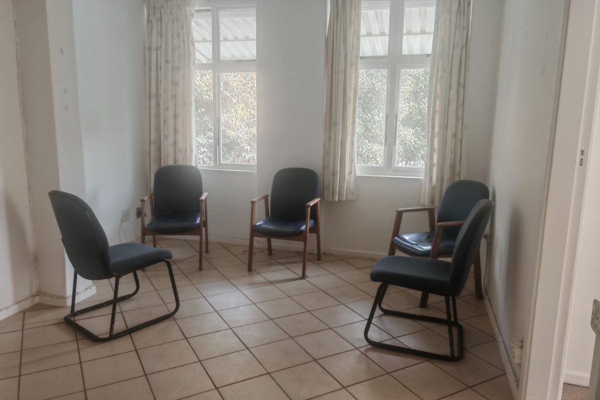 A living area featuring tiled flooring and dual casement windows, currently used as a seating space with dated wall finishes.