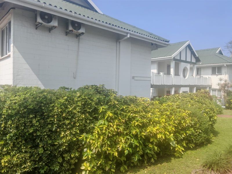 Exterior view of the apartment building featuring white facade, pitched roof, and balcony, surrounded by established garden landscaping.
