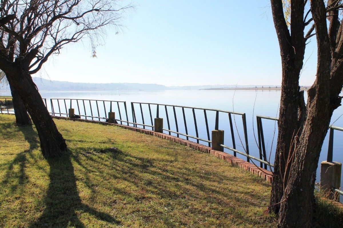 A scenic waterfront view featuring a grassy bank, a brick retaining wall, and a damaged metal safety railing overlooking the calm lake.