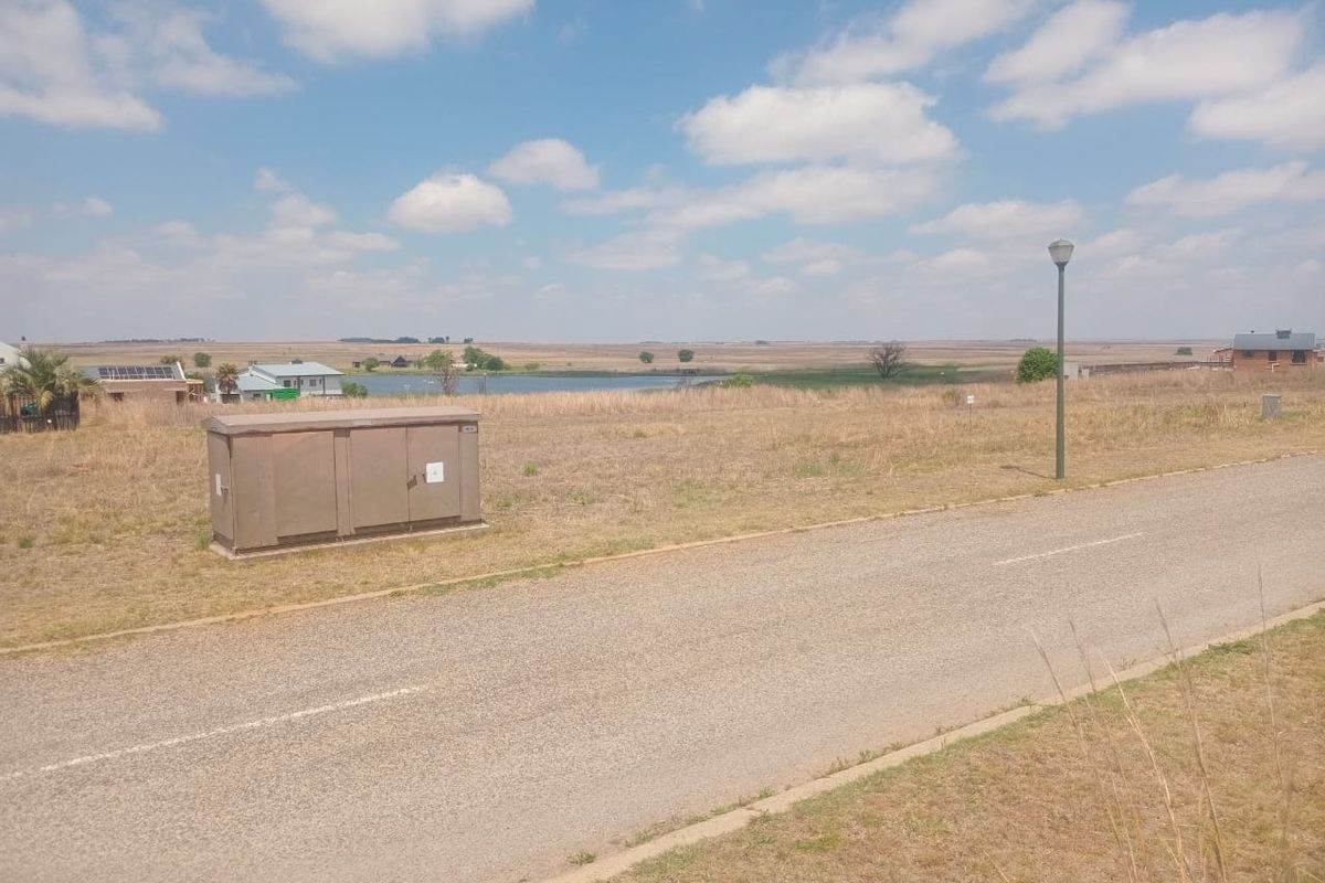 A view of a vacant residential plot featuring a paved road, street lighting, and an electrical utility box with surrounding landscape.