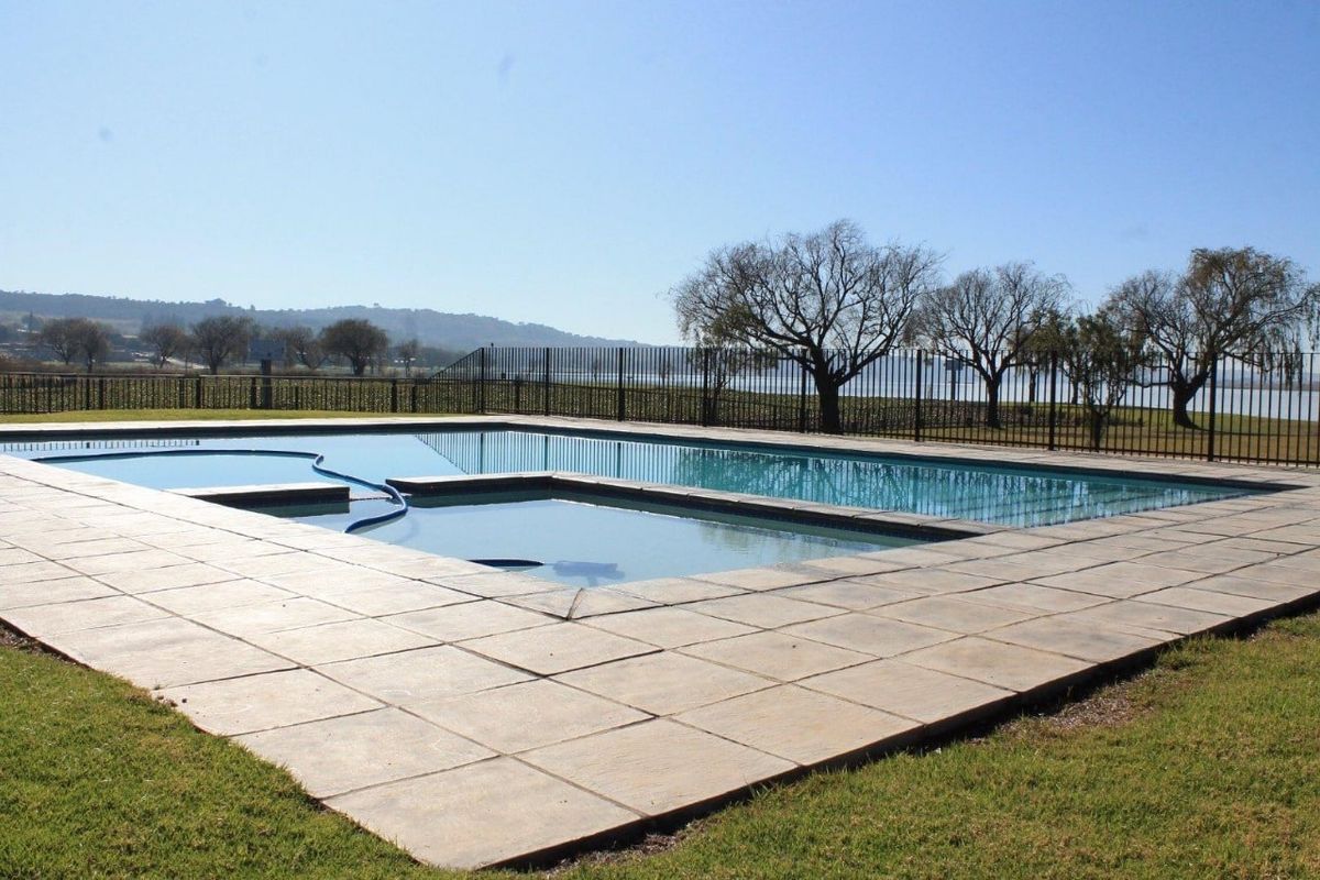 A well-maintained swimming pool area featuring a paved deck and metal perimeter fencing, set against a scenic landscape background.