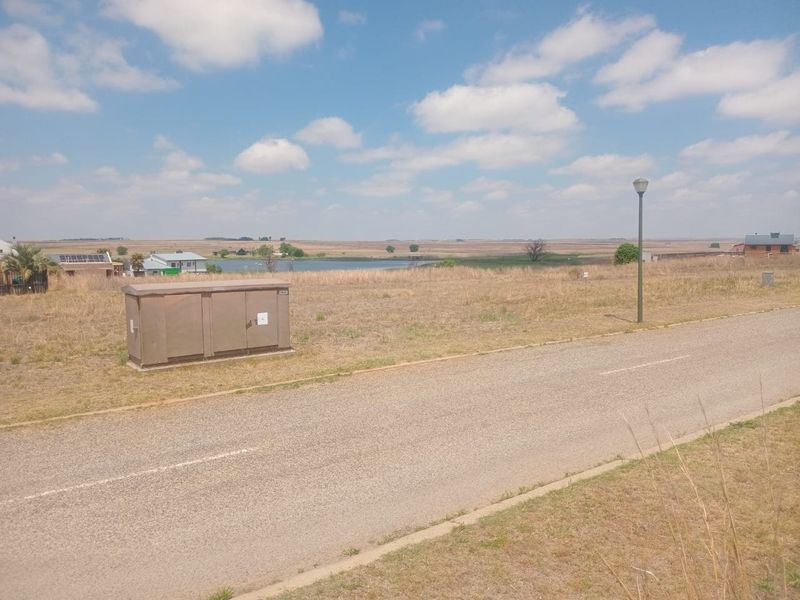 A view of a vacant residential plot featuring a paved road, street lighting, and an electrical utility box with surrounding landscape.