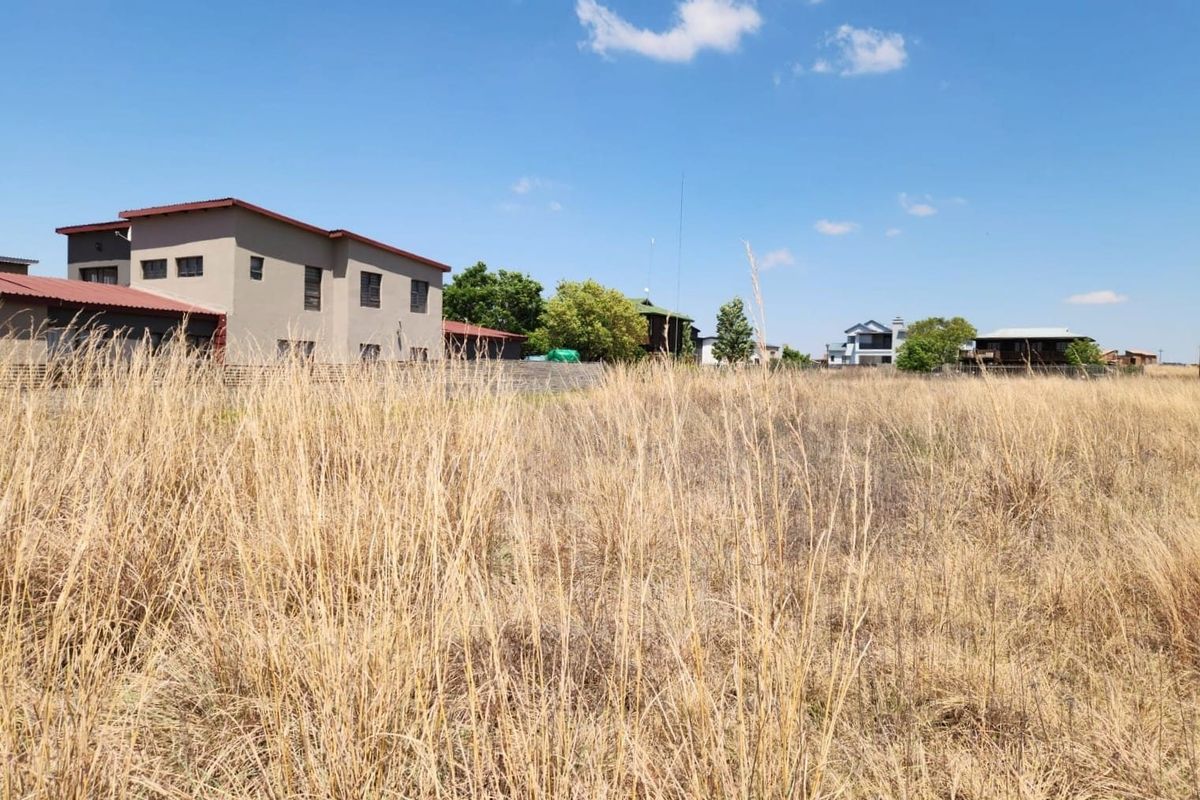 A view of a vacant residential plot featuring dry grass, with neighboring houses visible in the background.