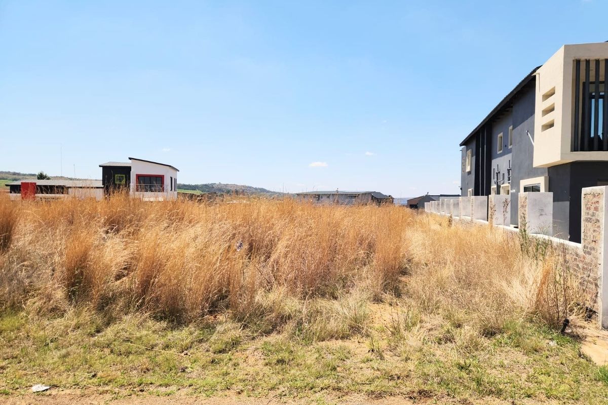 A vacant residential plot of land situated in a developed estate, featuring dry grass and adjacent modern residential properties.