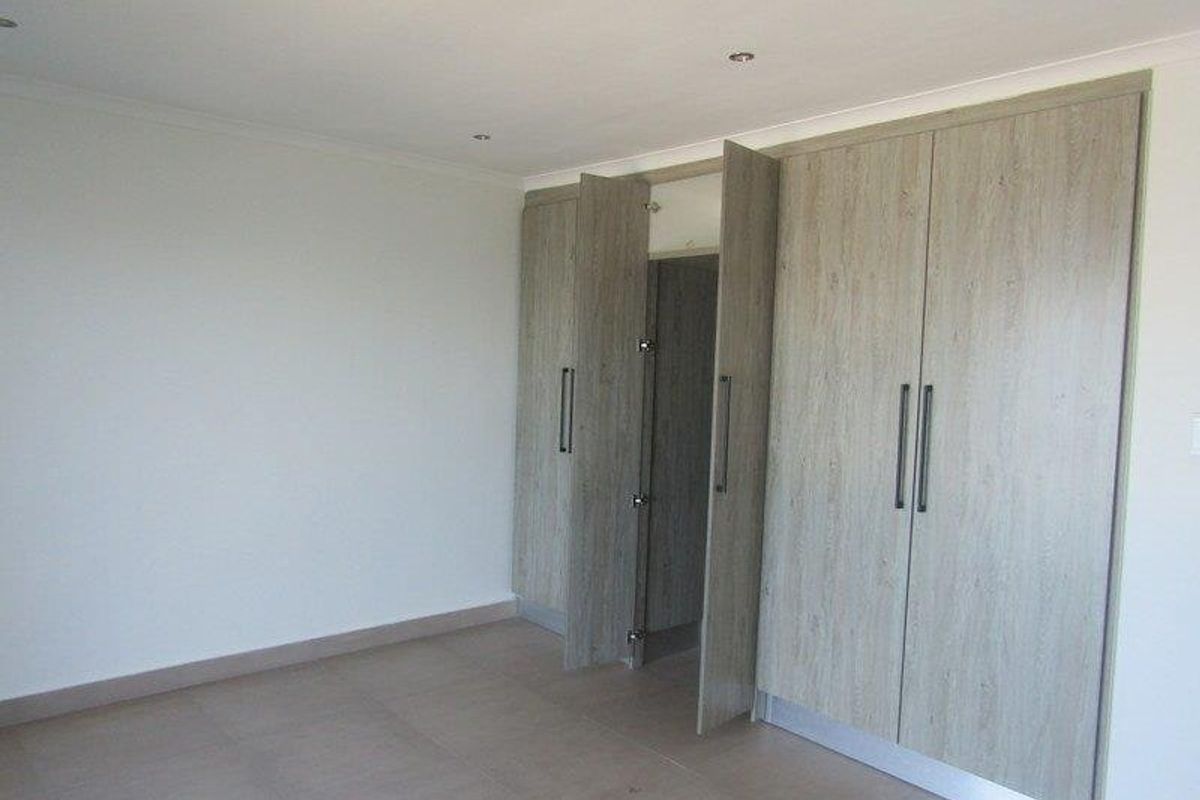 A bright, empty bedroom featuring modern wood-look built-in cupboards, neutral tiled flooring, and recessed ceiling lighting.