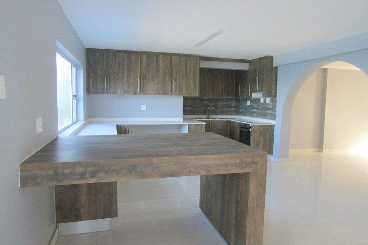 A clean, modern kitchen featuring wood-look cabinetry, a breakfast bar, tiled flooring, and an open-plan layout with an arched transition to the living area.