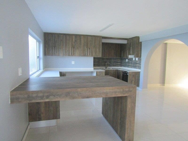 A clean, modern kitchen featuring wood-look cabinetry, a breakfast bar, tiled flooring, and an open-plan layout with an arched transition to the living area.