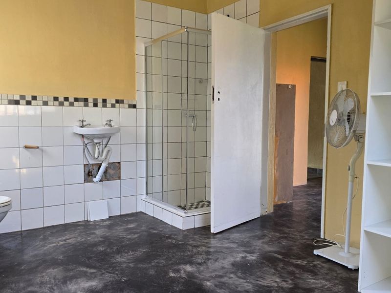 A functional bathroom featuring a shower enclosure, wall-mounted basin, and toilet, with some visible wear and exposed plumbing requiring maintenance.