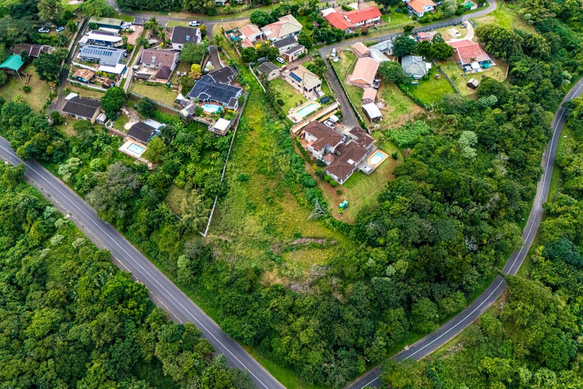 An aerial view of a vacant residential plot surrounded by established homes and lush greenery, situated at a road intersection.