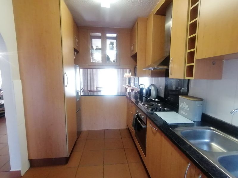 A functional galley-style kitchen featuring wood-finish cabinetry, granite countertops, built-in oven, gas hob, and tiled flooring, with a convenient pass-through window to the dining area.