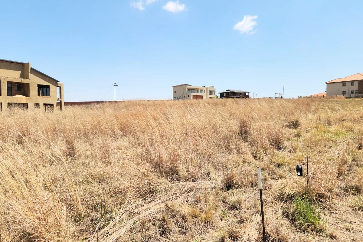 A vacant residential plot covered in dry grass, situated in a developed estate with surrounding residential properties.