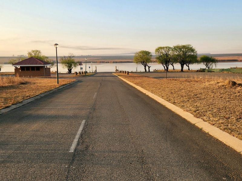 A paved access road leading towards a waterfront area, featuring a small brick structure and open landscape views.