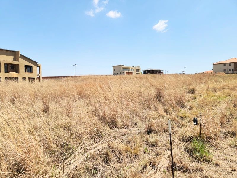 A vacant residential plot covered in dry grass, situated in a developed estate with surrounding residential properties.