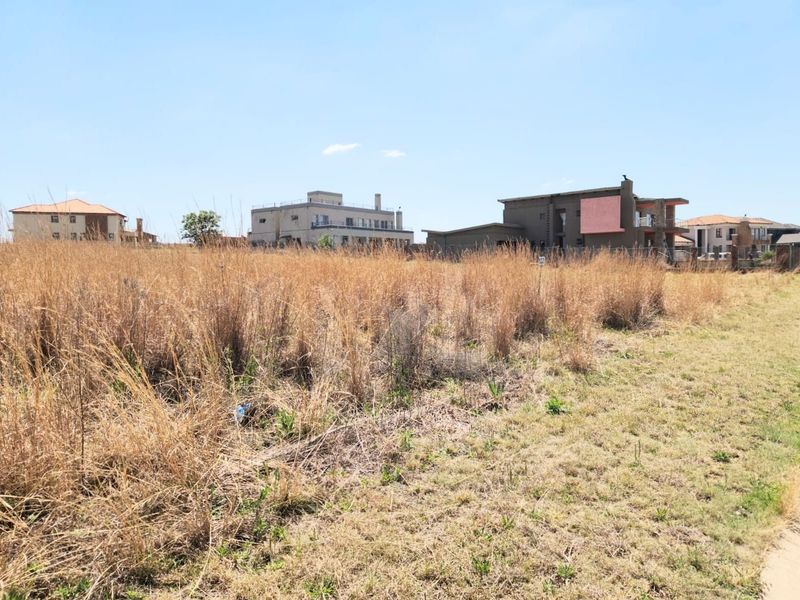 A vacant residential plot featuring dry grass and surrounding established residential properties in a developed neighborhood.