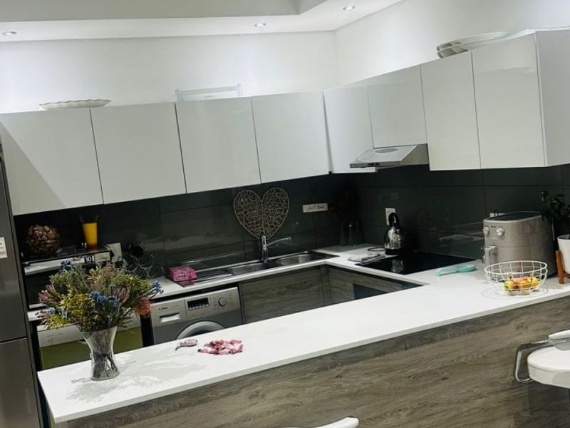 Modern kitchen featuring white gloss cabinetry, wood-look breakfast bar, stone-look countertops, dark glass splashback, and recessed downlighting.