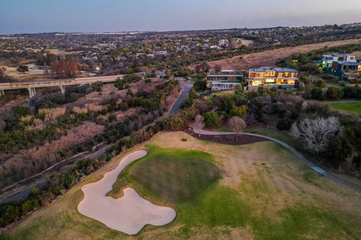 Aerial view of a golf course green and sand bunker, with residential properties and a bridge visible in the surrounding landscape.
