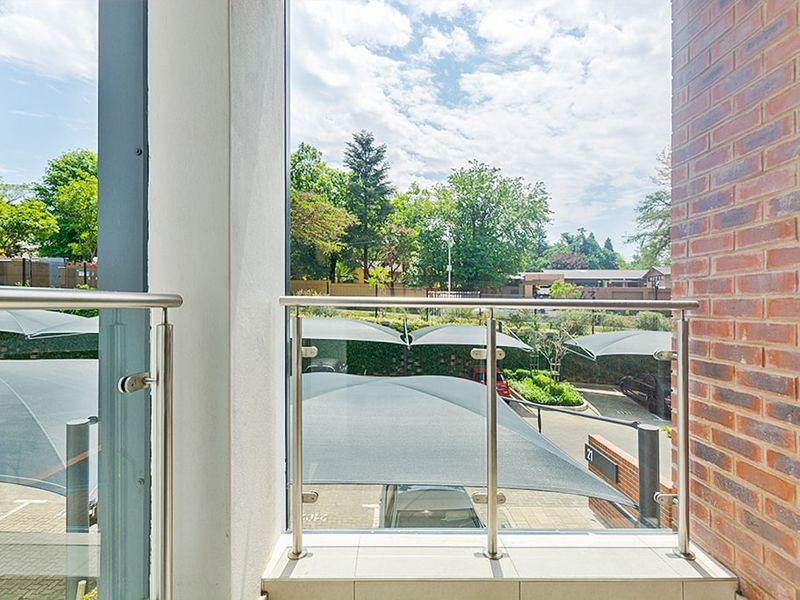 A modern balcony featuring a stainless steel and glass balustrade, tiled flooring, and exposed brickwork, overlooking the complex parking area.