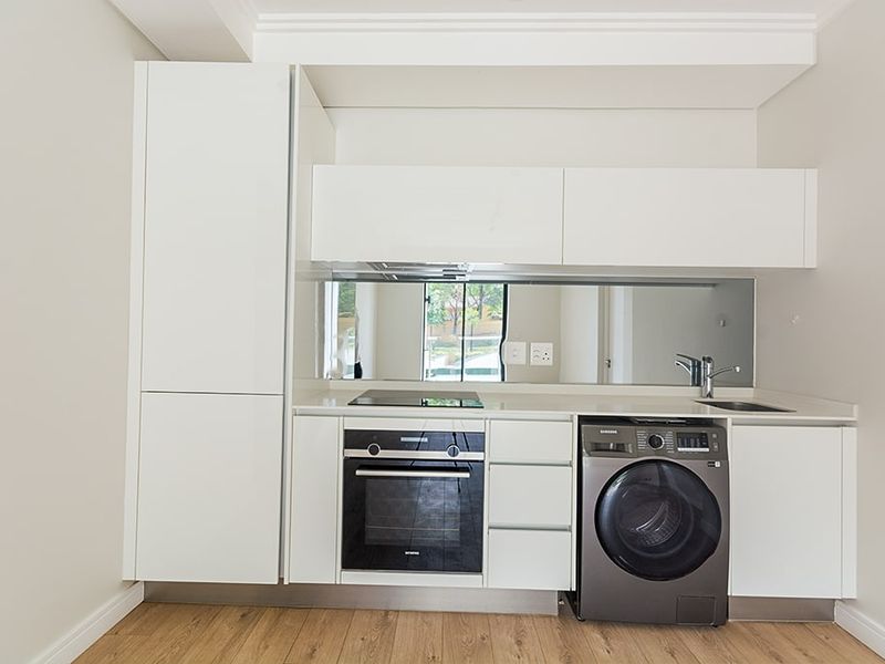 Modern compact kitchen featuring white cabinetry, integrated oven and hob, mirror splashback, and wood-look laminate flooring.