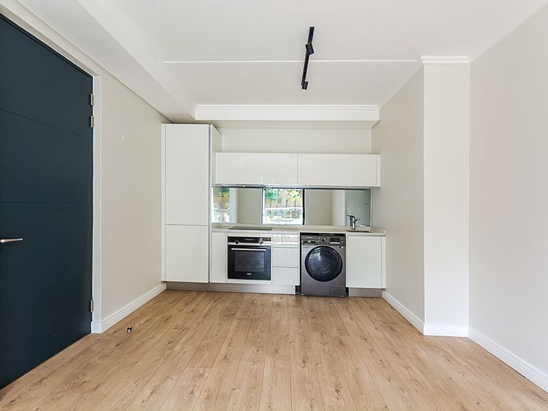 Modern compact kitchen featuring white cabinetry, integrated oven, hob, mirror splashback, and light wood-look laminate flooring in an open-plan living space.