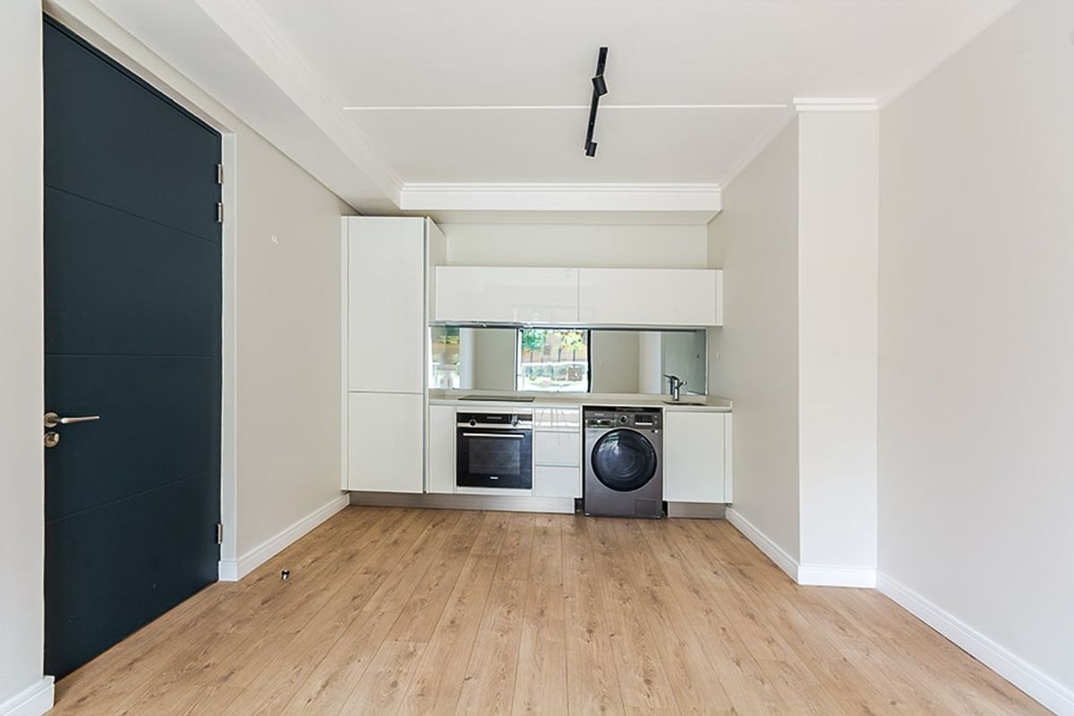 Modern compact kitchen featuring white cabinetry, integrated oven, hob, mirror splashback, and light wood-look laminate flooring in an open-plan living space.