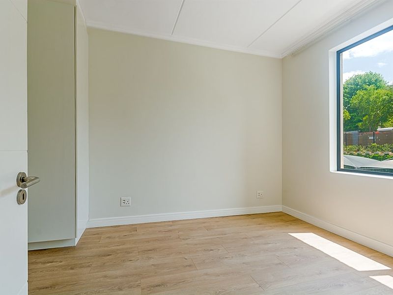 A bright, empty bedroom featuring modern laminate flooring, neutral walls, and a built-in cupboard with natural light from the window.