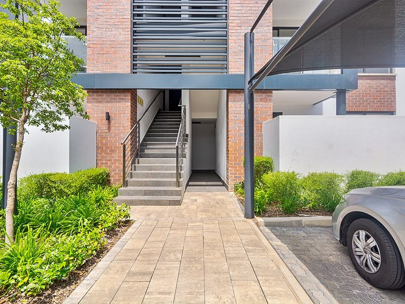 Modern apartment building entrance featuring a brick facade, paved walkway, metal staircase, and integrated shade sail for covered parking.