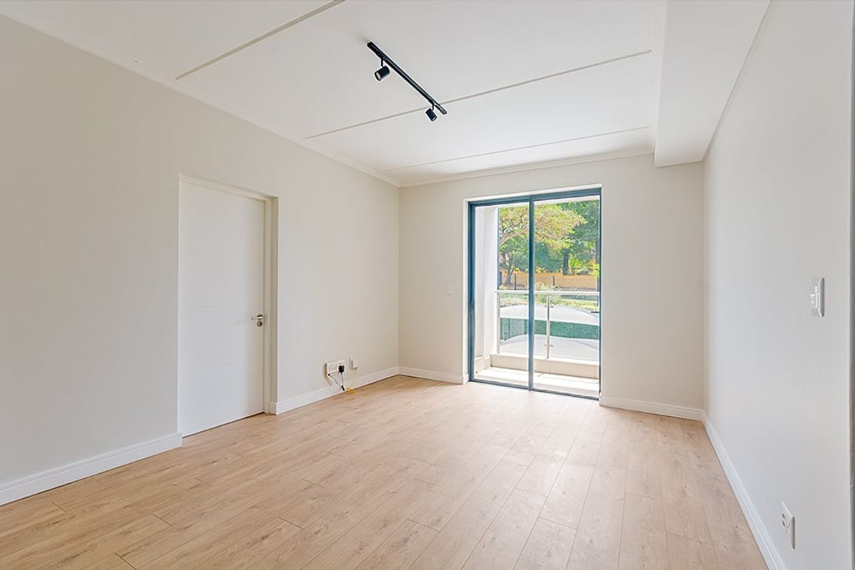 A bright, empty living room featuring modern laminate flooring, track lighting, and a sliding glass door providing access to a private balcony.