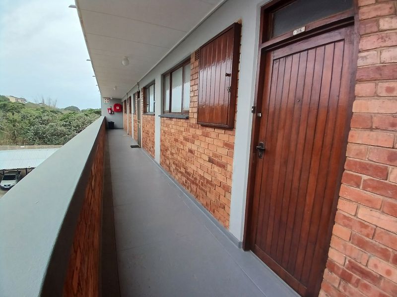 An exterior apartment building walkway featuring exposed brick walls, timber entrance doors, and a protective balustrade overlooking the parking area.