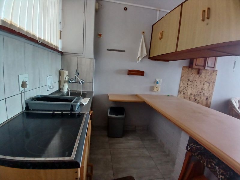 A dated kitchen featuring built-in cabinetry, an electric hob, a stainless steel sink, and a breakfast bar area with tiled flooring.